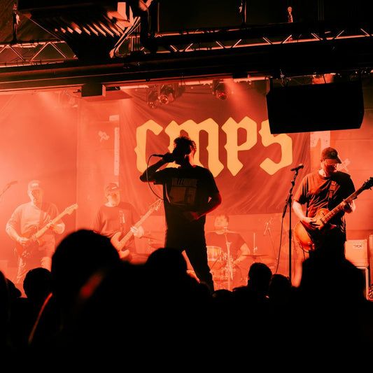 A band performing on stage with a backdrop featuring their band name in bold letters. The backdrop is displayed in a dimly lit environment during a concert.