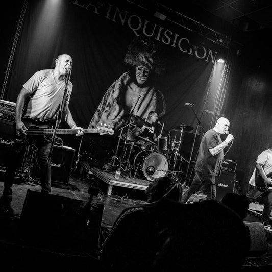 A black and white photo of a band performing on stage with a backdrop that has a design and the text 'LA INQUISICION'.